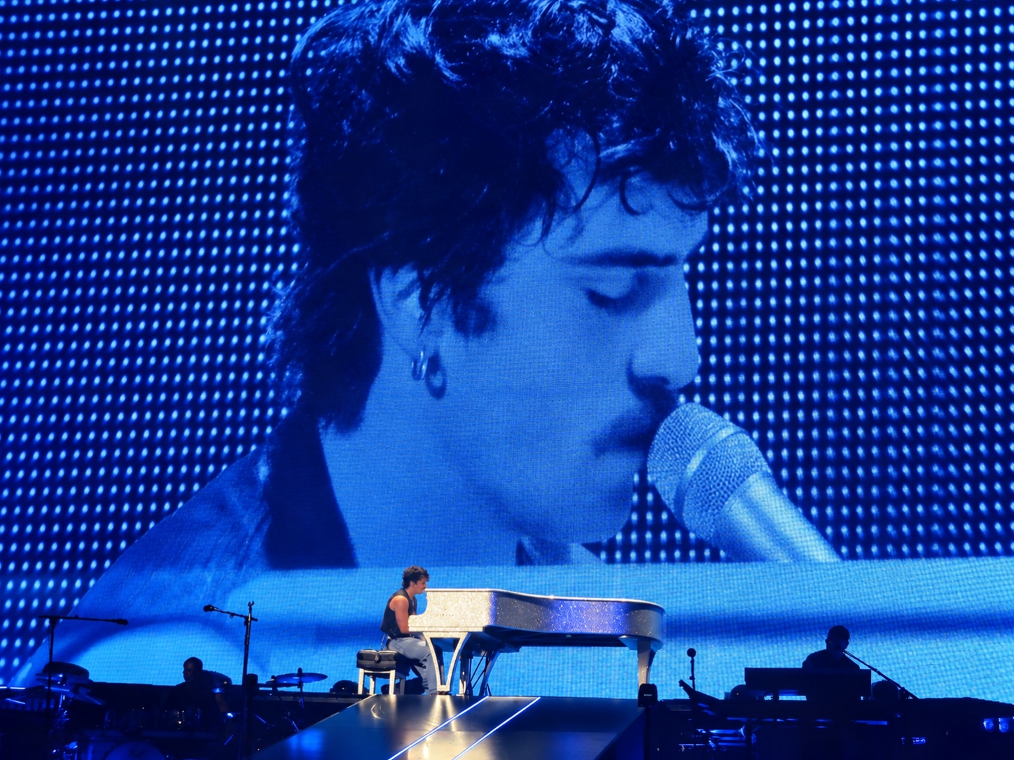Wide concert photograph of a singer at a grand piano with a blue screen behind the stage