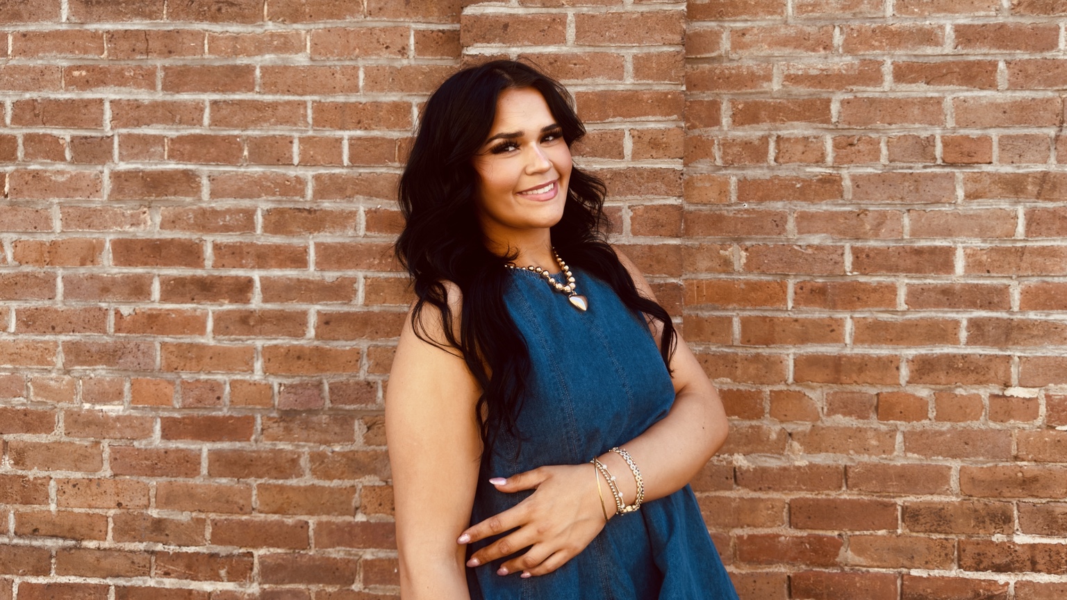 Smiling woman in a denim dress with warm brickwork behind her